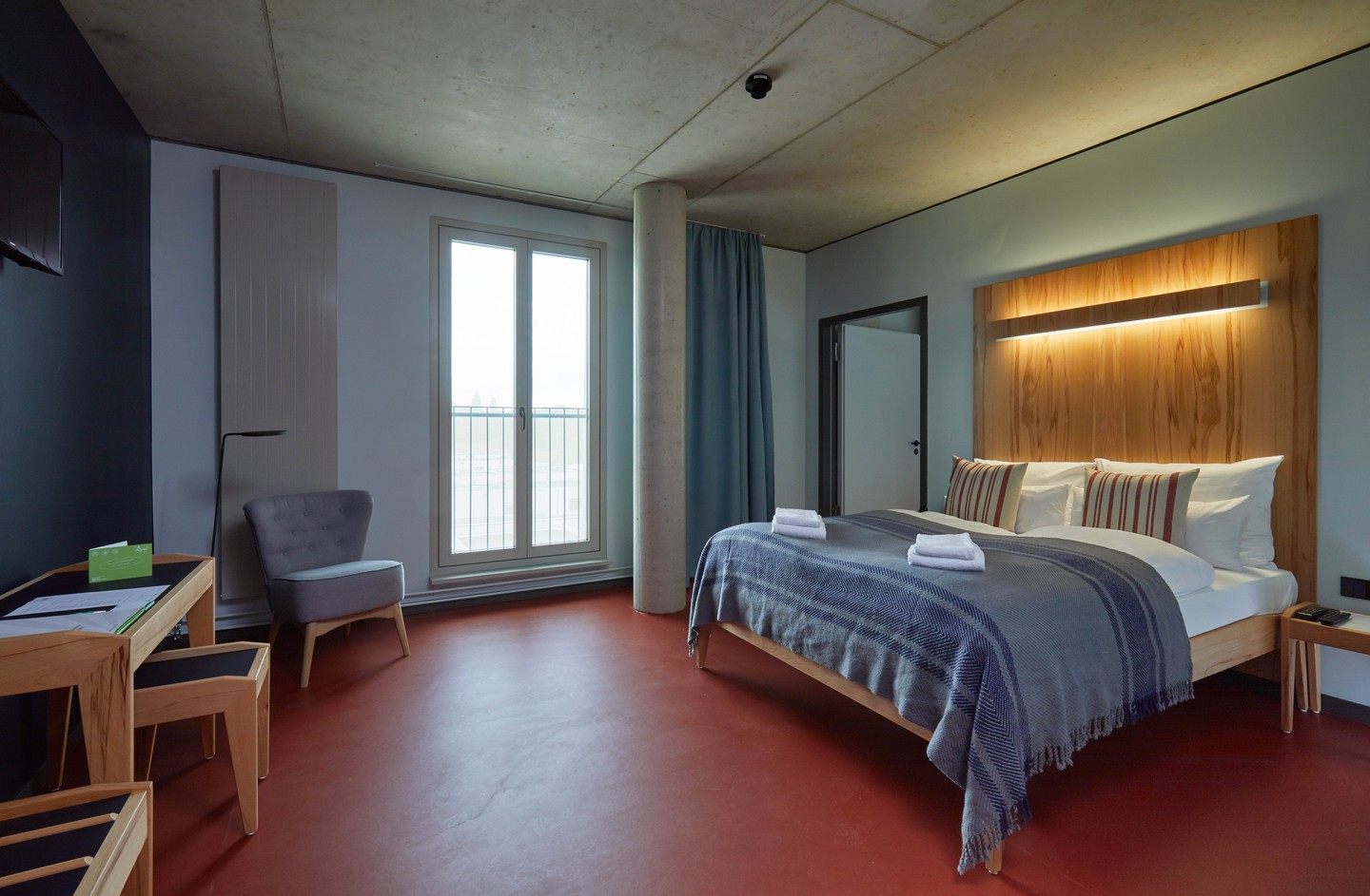 Modern eingerichtetes Zimmer mit großem schlichten Holzbett. Das Design wird bestimmt durch klare Linien aus Holz, Beton und Blau- und Rottönen. 
				