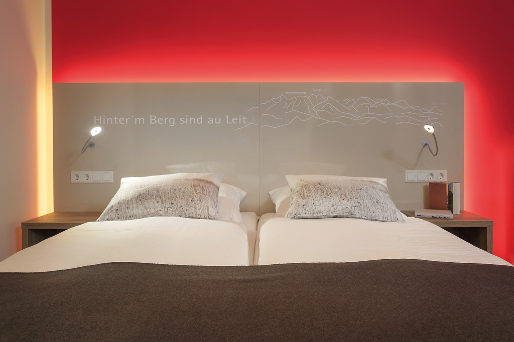 Doppelbett vor einer roten Wand mit den Allgäuer Bergen auf dem Kopfteil.
				